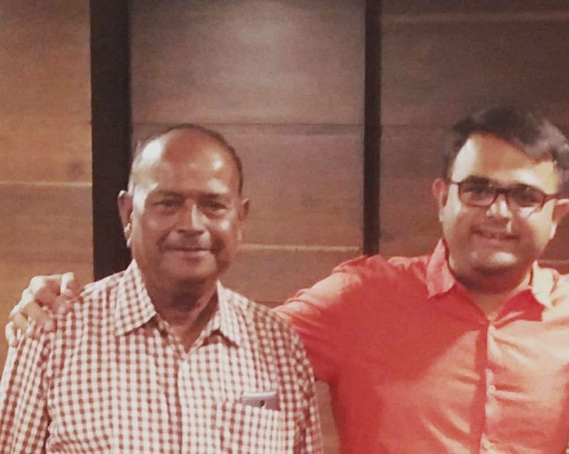 An older man in a checked shirt with his arm around a younger man in glasses and an orange shirt