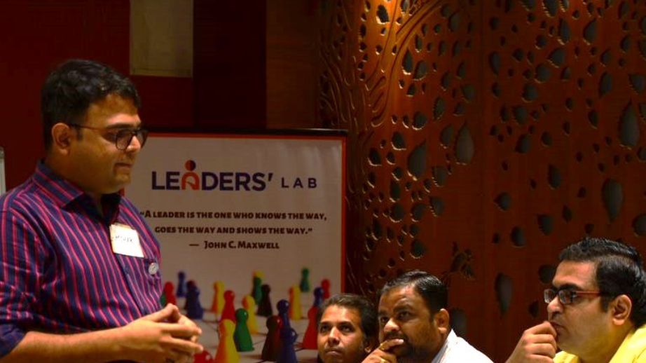 A speaker standing and talking to people seated at a table, with a Leaders' Lab banner behind them
