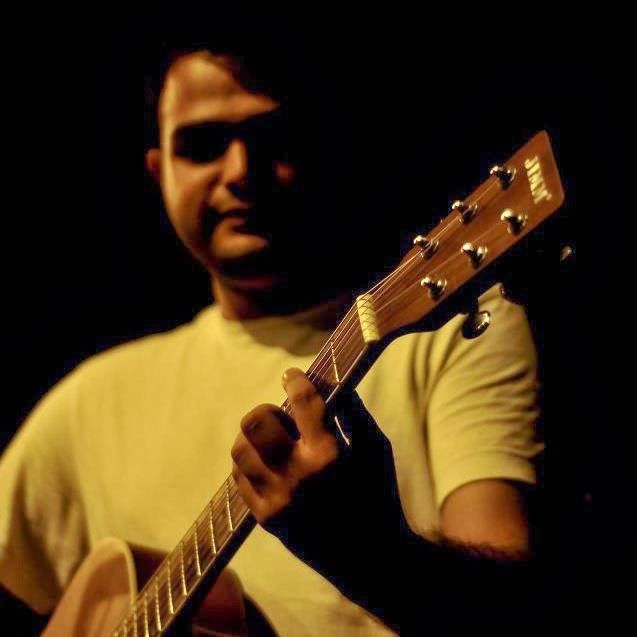 A man in a pale shirt playing an acoustic guitar, lit warmly against a dark background