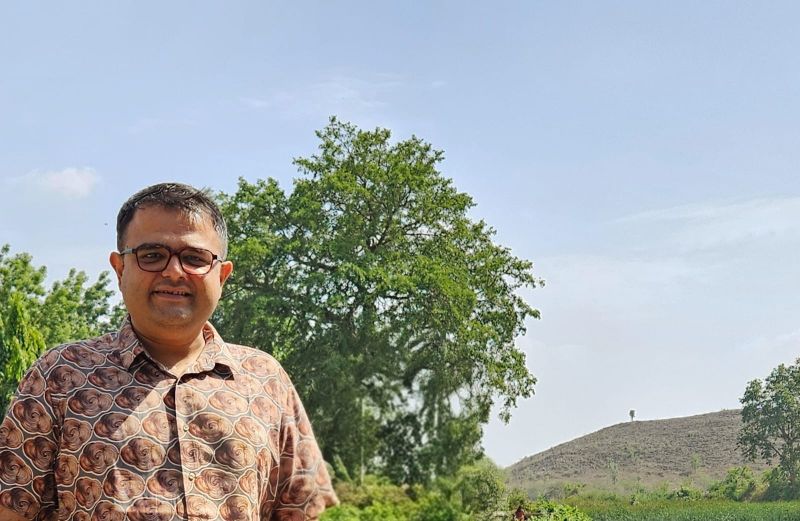 A smiling man in glasses and a patterned shirt standing in front of a large tree and a hill